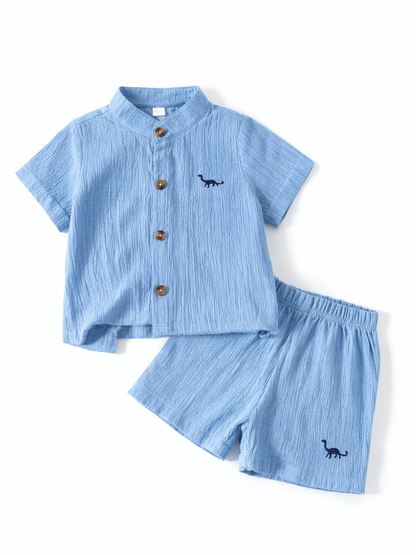 白背景に平置きされたキッズ用上下セット。シワ感のあるライトブルーの半袖シャツ（スタンドカラー、木目ボタン）と、同素材のウエストゴムショートパンツが同じ配置で並べられている。シャツとパンツにネイビーの恐竜刺繍が入っており、やわらかな影で立体感がある。
