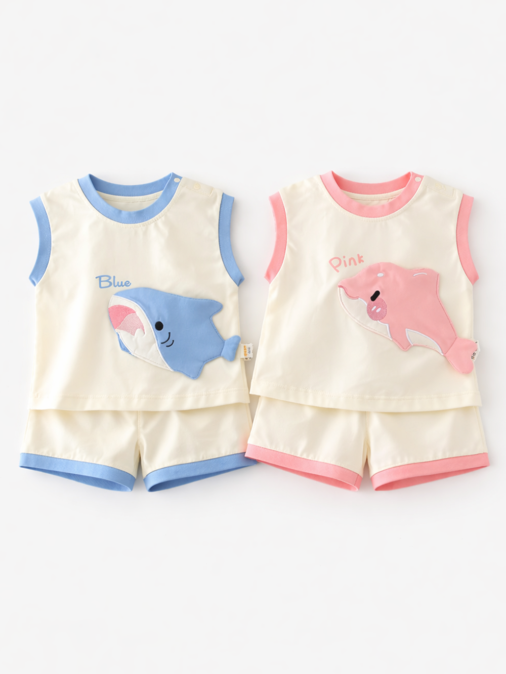 白背景に平置きされたベビー用ノースリーブ上下セット2組。左はブルーの縁取りで、サメのアップリケと「Blue」文字があるトップスと同色トリムのショートパンツ。右はピンクの縁取りで、イルカのアップリケと「Pink」文字があるトップスと同色トリムのショートパンツが並べられている。
