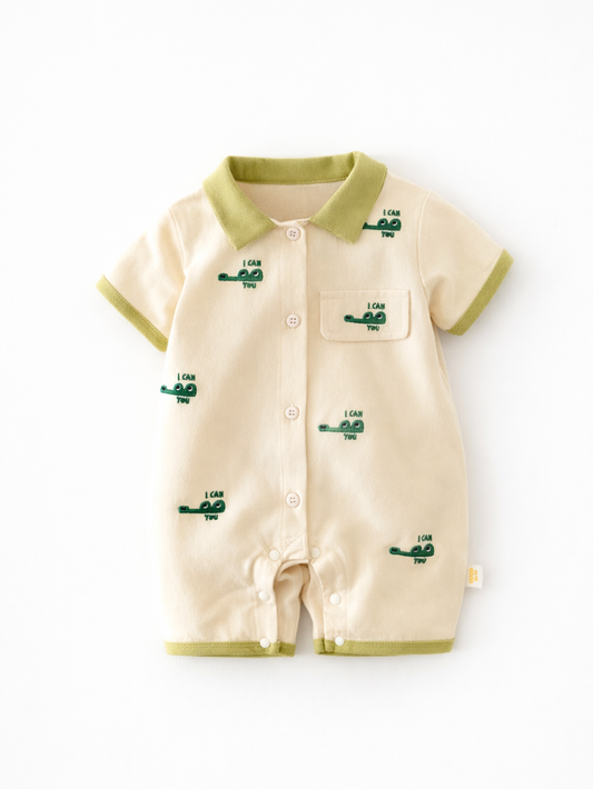 白背景に平置きされた、クリーム色のベビー用半袖ロンパース。グリーンの襟・袖口・裾のパイピングと、胸ポケット付き。全体に小さな車の刺繍が散りばめられている。
