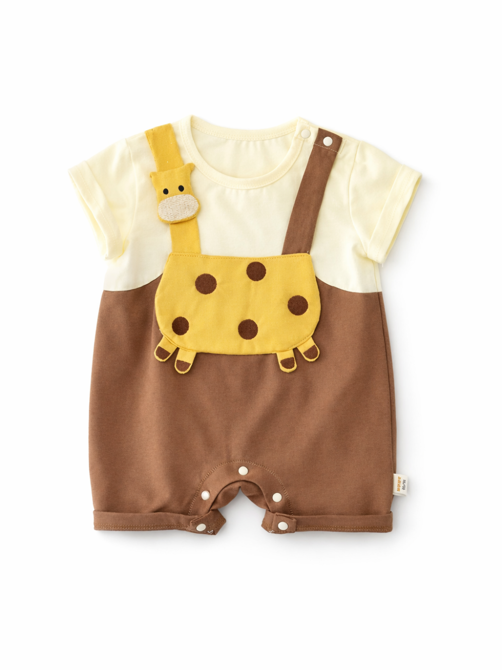 キリンモチーフのベビーロンパースを白背景でフラットレイ撮影。アイボリーの半袖トップにブラウンのサロペット風デザイン、イエローのドット柄ポケットとキリン顔のストラップ飾り、股下スナップボタン付きで、生地の質感と縫製がはっきり見える。