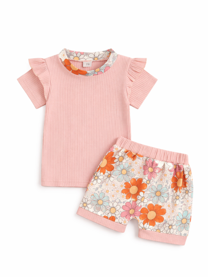 ベビー用の半袖トップスとショートパンツの2点セット。淡いピンクのリブ素材トップスはフリル袖で、花柄の襟ぐりデザイン。ショーツはレトロ調の花柄プリント（オレンジ・ピンク・ブルー系）で、ウエストと裾にピンクのリブ切替。白背景のスタジオ撮影で上下を並べて見せている。
