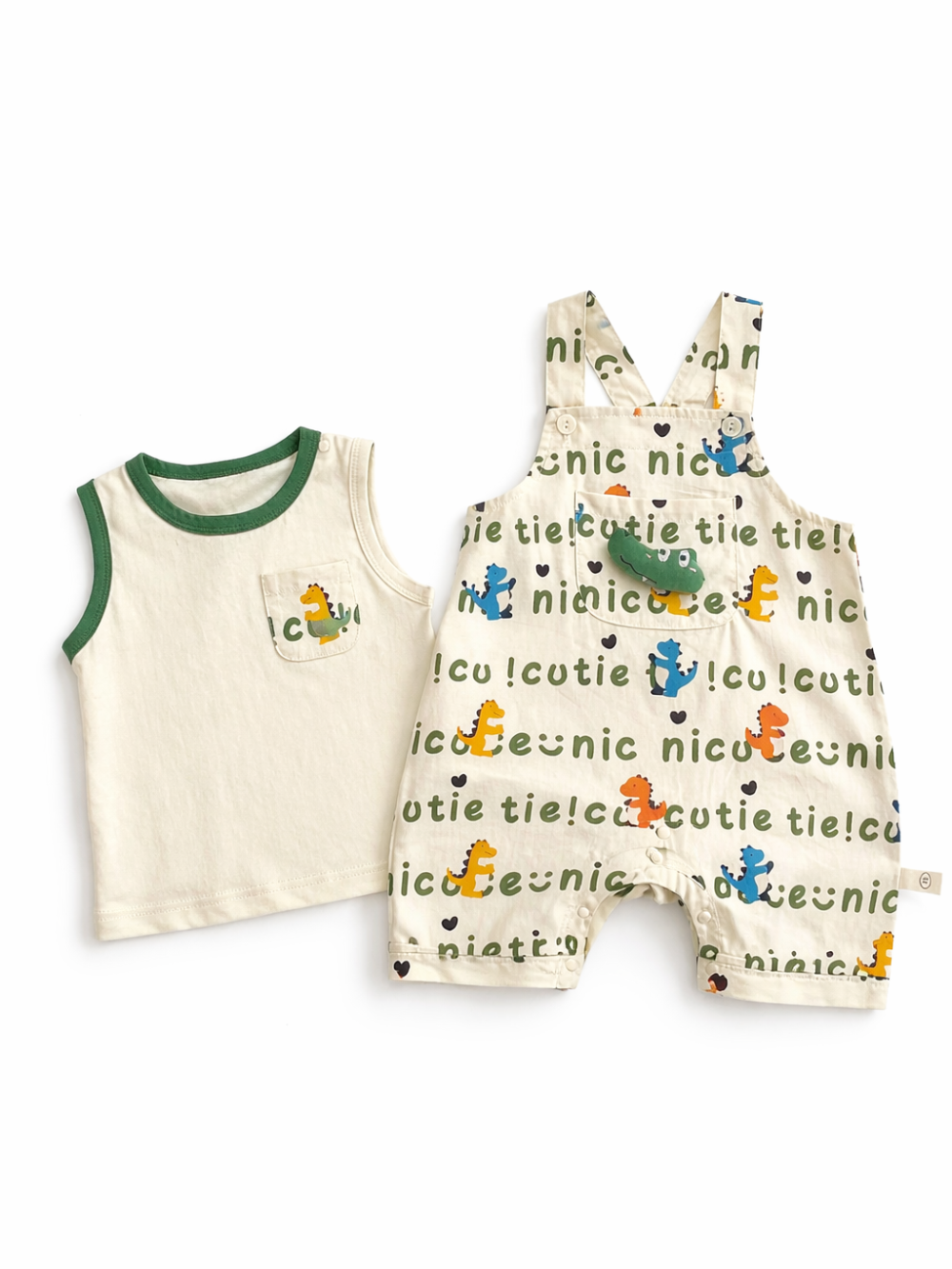 ベビー用のタンクトップとサロペットロンパースの2点セット。アイボリーのタンクトップはグリーンのパイピングで、胸ポケットに恐竜モチーフ付き。サロペットは「nice」「cutie」などの文字とカラフルな恐竜柄の総柄で、肩ひも付き・股下スナップ仕様。白背景のスタジオ撮影で2点を並べて見せている。
