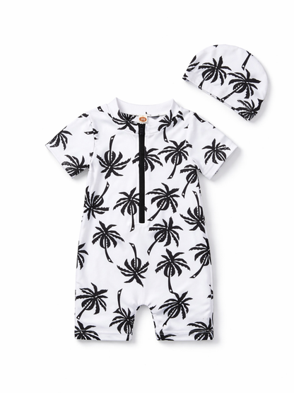 ベビー用の半袖ロンパース水着とスイムキャップの2点セット。白地に黒のヤシの木プリントで、前面に黒いハーフジップ付き。おそろい柄のキャップが付属。白背景のスタジオ撮影で上下を並べて見せている。
