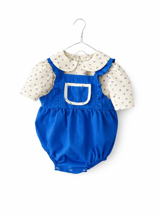 白背景で正面から撮影したベビー用ロンパース。ロイヤルブルーの吊りパンツ風デザインに、クリーム地の小花柄パフ袖ブラウスを重ねた2点風スタイルで、胸ポケットと股下スナップが見える。
