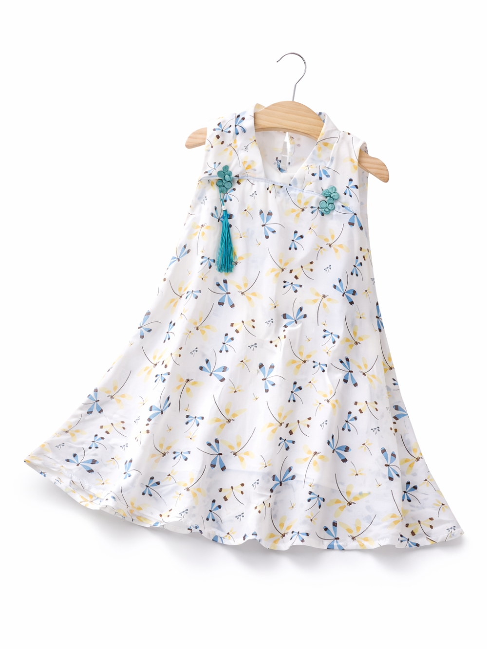 白地に青と黄色のトンボ柄が散りばめられた、ノースリーブの子ども用Aラインワンピース。襟元に青緑の飾りボタンとタッセルが付いており、木製ハンガーに掛けられて白背景で撮影されている。
