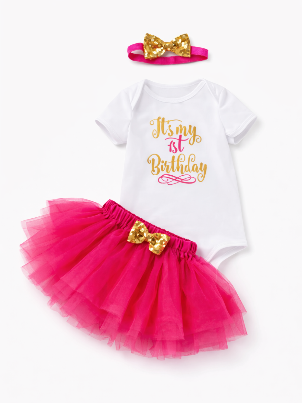純白（#FFFFFF）のスタジオ背景に、ベビー用の3点セットが平置きで配置されている。白い半袖ロンパースの中央に「It’s my 1st Birthday」のプリントがあり、濃いピンクのチュチュスカートにはウエスト部分にゴールドのスパンコールリボンが付いている。上部には同じゴールドリボン付きのヘアバンドが置かれ、全体が明るく均一な光でくっきり写っている。
