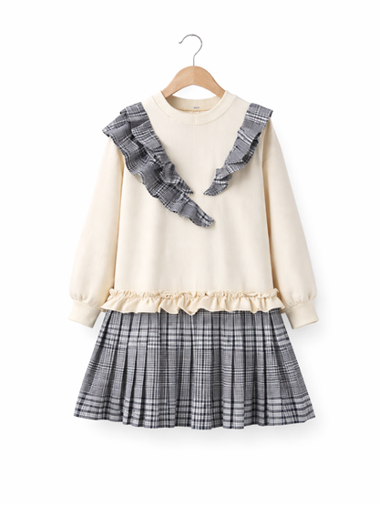 純白（#FFFFFF）のスタジオ背景に、木製ハンガーに掛けられた子ども用ワンピースが正面から写っている。クリーム色の長袖トップに、ネイビー×グレー系チェックのV字フリルが胸元に付いており、ウエストにはクリーム色のフリル切り替え、下部は同柄のプリーツスカートになっている。均一な照明で質感と柄がくっきり見え、下に自然な微影が入って立体感がある。
