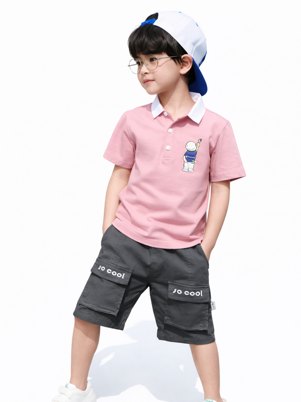 白背景で撮影したキッズモデル着用画像。ピンクの半袖ポロシャツ（胸元にキャラクターワンポイント）と、グレーのカーゴショーツ（ポケットに「so cool」プリント）を着用し、後ろ向きキャップとメガネを合わせたコーデ。
