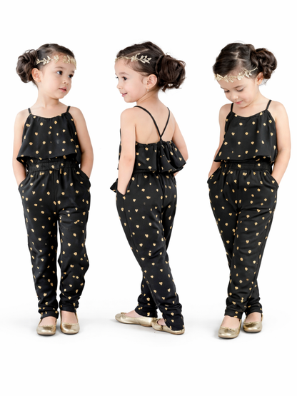 白背景のスタジオで、黒地にゴールドのハート柄が入ったキャミソールタイプのキッズオールインワンを着た女の子。正面・後ろ姿・うつむきの3ポーズでデザインを見せており、髪はお団子、ゴールドのヘッドアクセとゴールドのフラットシューズを合わせている。
