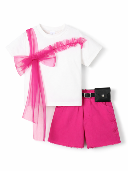 白背景のスタジオ撮影で、白い半袖Tシャツとショッキングピンクのショートパンツのキッズ2点セット。Tシャツにはピンクのチュールリボンとフリル装飾が斜めに付いており、パンツはフリンジ裾で黒いベルトとミニポーチが付いている。
