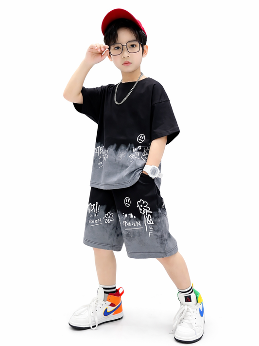 白背景のスタジオ撮影で、黒×グレーのグラデーションTシャツと総柄ショートパンツのストリート系キッズセットアップを着た男の子。赤いキャップとメガネ、チェーンネックレスを合わせ、ポーズを取りながら立っている。
