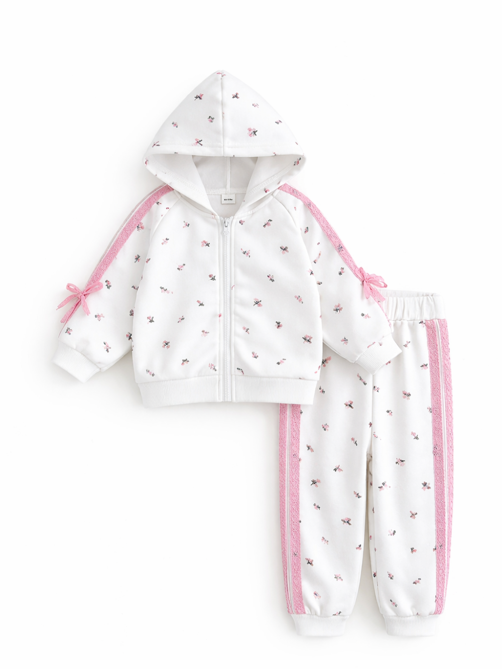 白背景のスタジオ撮影で、小花柄の白いキッズセットアップ。フード付きジップパーカーとジョガーパンツの2点セットで、袖とパンツ脇にピンクのレース風ラインが入っている。

