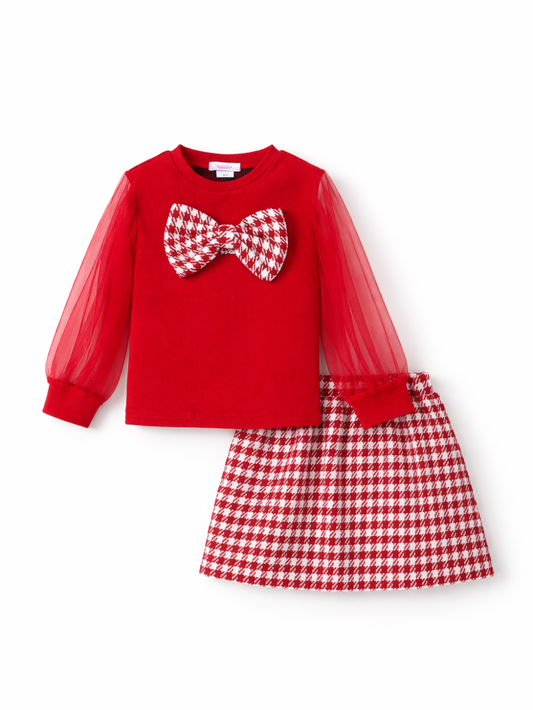 白背景のスタジオ撮影で、赤い長袖トップスと千鳥格子柄スカートのキッズ2点セット。トップスはシアーなチュール袖と胸元の大きなリボンが特徴で、スカートは同柄の台形シルエットになっている。
