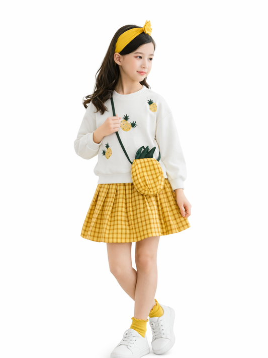 白背景のスタジオ撮影で、パイナップル刺繍の白いスウェットとイエローのギンガムチェック柄スカートを着た女の子が、片足を軽くクロスしたカタログ風ポーズで立っている。黄色のヘアバンドとパイナップル型ポシェット、白いスニーカー、黄色のソックスを合わせている。
