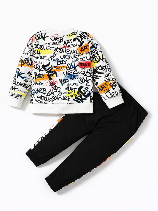 カラフルなグラフィティ風ロゴ総柄のキッズ長袖スウェットと、ブラックのジョガーパンツの上下2点セット。パンツ脇に同柄の切り替えライン入り。白背景のスタジオ撮影。
