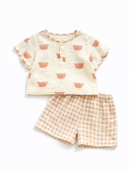 くま顔プリントのキッズ上下2点セット。アイボリーの凹凸素材半袖トップス（前ボタン付き）と、オレンジ×アイボリーのギンガムチェックショートパンツの組み合わせ。白背景のスタジオ撮影。
