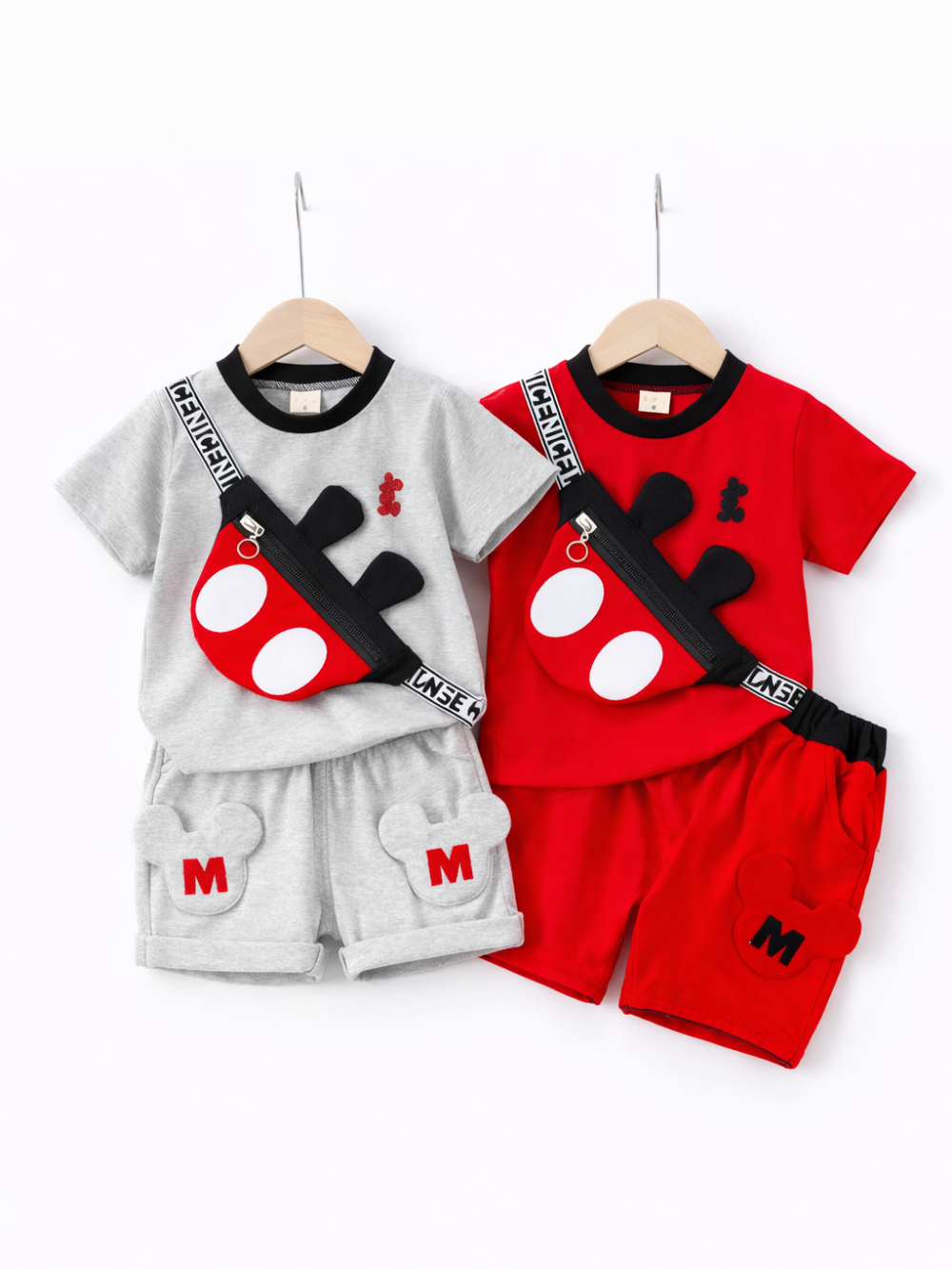 キッズ半袖上下セット2色（グレー／レッド）。黒リブ襟のTシャツとショートパンツのセットで、パンツに耳型ワッペンと「M」刺繍入り。赤×黒の斜め掛けミニバッグ付き（耳モチーフ・ロゴ入りストラップ）。白背景のスタジオ撮影。
