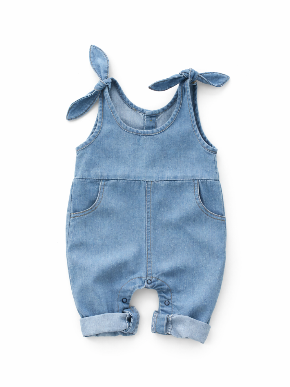 白背景のスタジオ撮影で、淡いブルーデニムのベビーロンパースが平置きで写っている。肩ひもはリボン結びで、前ポケットと股下スナップ、裾のロールアップが見える。
