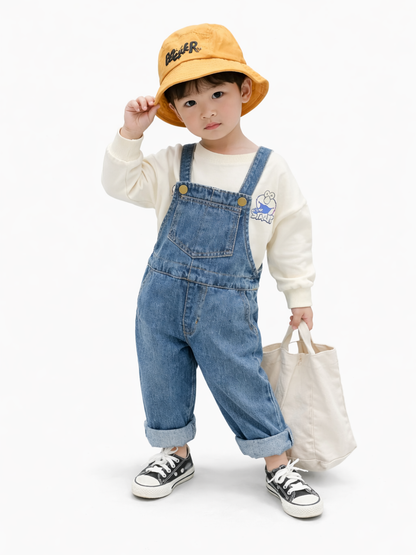 白背景のスタジオ撮影で、男の子モデルがデニムのオーバーオールとクリーム色のスウェットを着用し、黄色のバケットハットをかぶって立っている。片手で帽子に触れ、もう片手で生成りのトートバッグを持ち、黒白スニーカーが見える。
