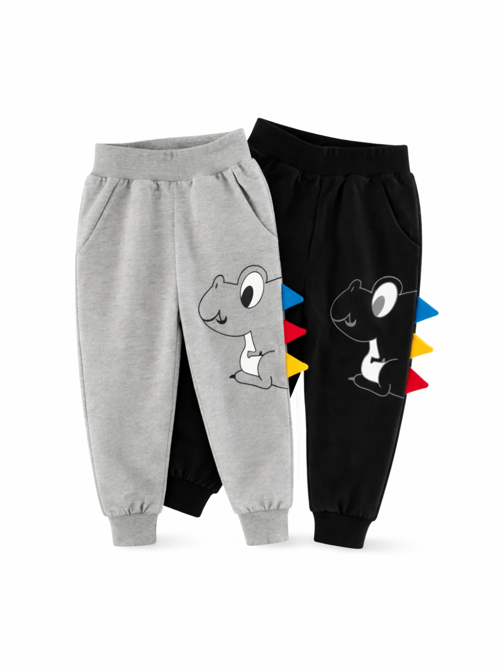 白背景のスタジオ撮影で、キッズ用ジョガーパンツが2本並んで写っている。左はグレー、右はブラックで、恐竜のイラストプリントとサイドに赤・青・黄の立体スパイク装飾、ウエストと裾はゴム仕様。
