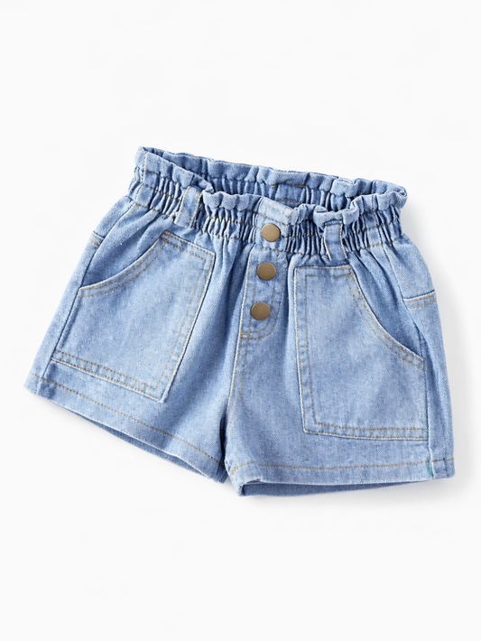 白背景のスタジオ撮影で、ライトブルーのキッズデニムショートパンツが平置きで写っている。フリル状のゴムウエスト、フロントの3つボタン、大きめの前ポケット、裾のステッチが見える。
