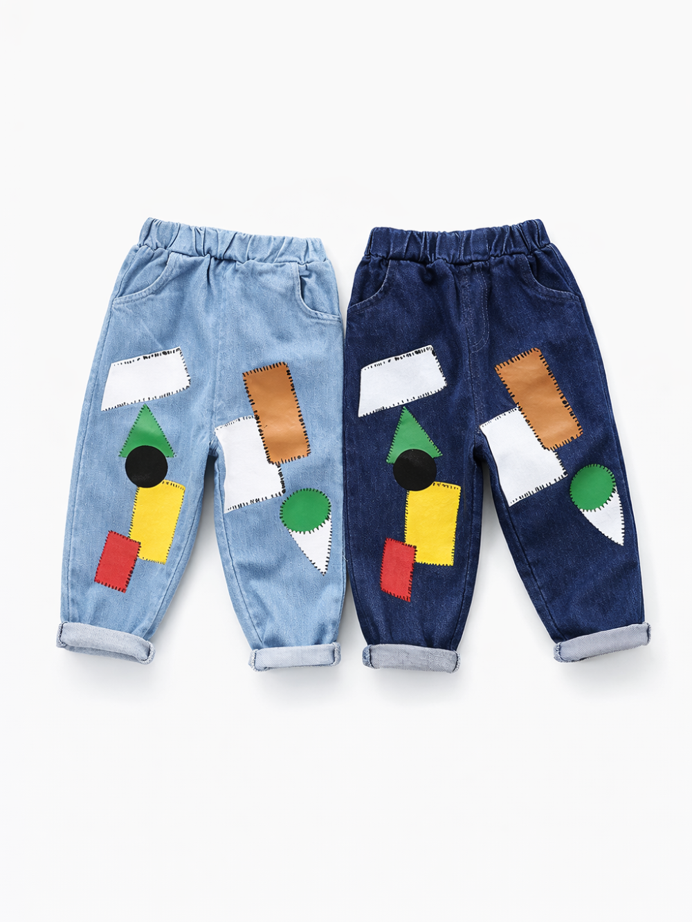 白背景のスタジオ撮影で、キッズ用デニムパンツが2本並んで平置きされている。左はライトブルー、右は濃紺で、どちらもウエストゴム仕様と裾のロールアップがあり、前面に白・茶・緑・黄・赤・黒の幾何学パッチが縫い付けられている。
