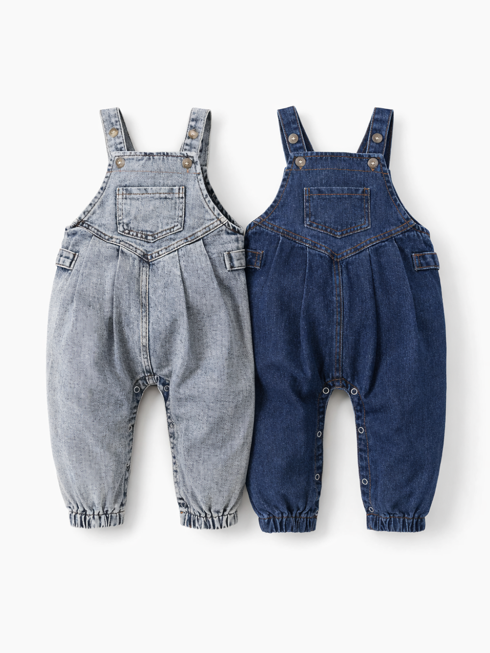 白背景のスタジオ撮影で、ベビー用デニムサロペットが2点並べて平置きされている。左はウォッシュ加工のライトグレー系デニム、右は濃紺デニムで、どちらも肩ストラップのボタン留め、胸ポケット、股下スナップ、裾ゴム仕様が見える。
