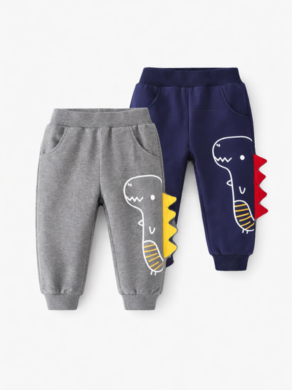 白背景のスタジオ撮影で、キッズ用ジョガーパンツが2本並んで写っている。手前はグレー、奥はネイビーで、どちらも恐竜のプリントとサイドの立体スパイク装飾（グレーは黄色、ネイビーは赤）、ウエストと裾はゴム仕様、前ポケット付き。
