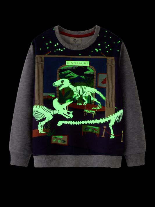 黒に近い暗背景の正面写真。グレー袖の子ども用スウェット。フロントに恐竜博物館風プリントで、恐竜の骨格と星柄が黄緑に発光して見える（蓄光デザイン）。
