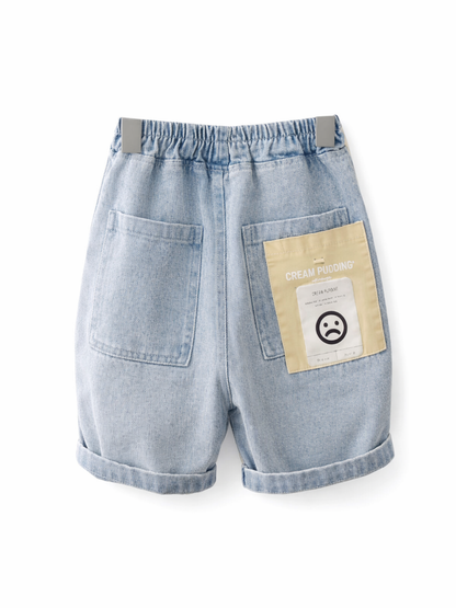 ライトブルーのデニムショートパンツ（バックスタイル）。ウエストはゴム仕様で、背面に大きめポケット2つ、右ポケットにクリーム色のラベルパッチと顔アイコンデザイン。裾は折り返し。白背景の商品単体画像。
