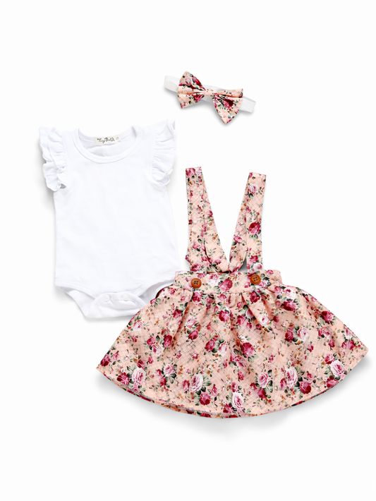 ベビー女の子用3点セット。ホワイトのフリル袖ロンパース、ピンク系フローラル柄のサスペンダースカート（木ボタン付き）、同柄のリボンヘアバンドの組み合わせ。白背景の商品画像。
