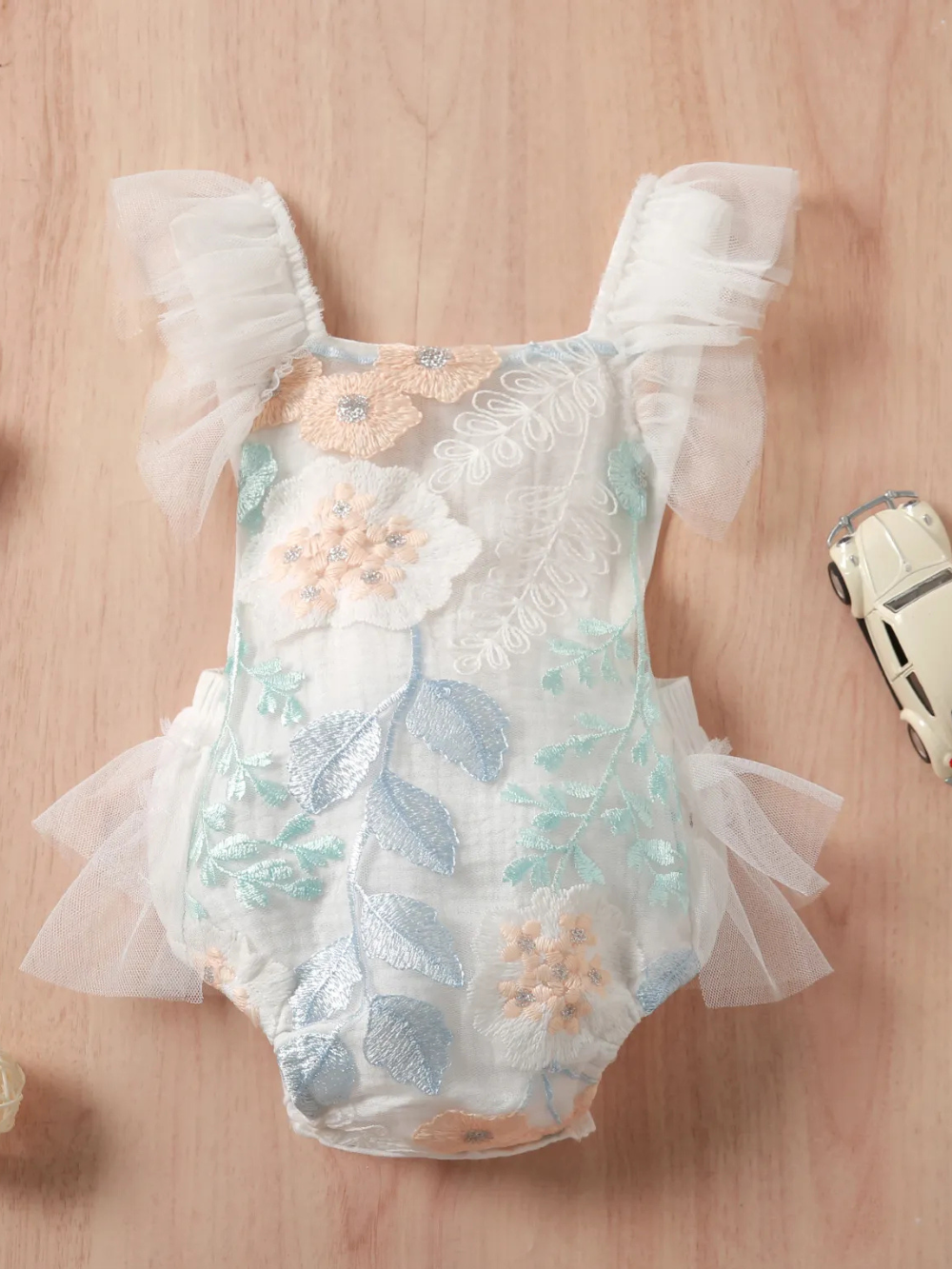 ベビー用チュール付きロンパース。ホワイトベースにパステルカラーの花柄刺繍（ピーチ、ミント、ライトブルー）とシルバー調アクセント入り。肩はチュールのフリルストラップ、サイドにもチュール飾り付き。白背景の商品単体画像。
