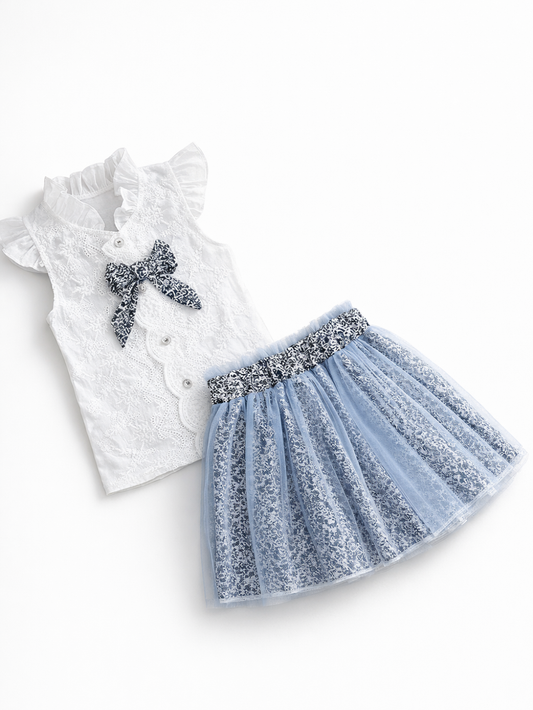 白いレースのフリル襟ノースリーブブラウス（花柄リボン付き）と、ブルーの花柄チュール重ねスカートの2点セット。上品で軽やかな女の子用コーデ。
