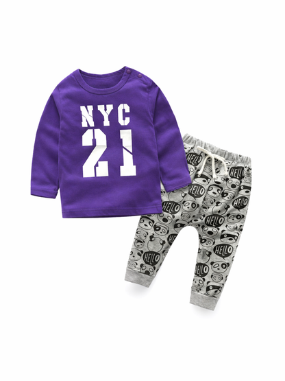 白いスタジオ背景で撮影した、ベビー／キッズ用2点セット。パープルの長袖トップスはフロントに「NYC 21」プリント、肩スナップ付き。ボトムはグレーの総柄ジョガーパンツで「HELLO」とキャラクター柄、ウエストはドローストリング仕様。セット全体が見えるカタログ写真。
