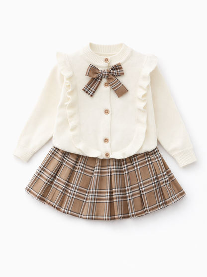 白いスタジオ背景で撮影した、女の子用トップス＋スカートの2点セット。アイボリーのニットカーディガンはフリル縁取りと木調ボタン付きで、首元にブラウンチェックのリボン。ボトムはブラウン系チェック柄のプリーツスカート。セット全体が見えるカタログ写真。
