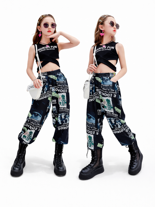 白いスタジオ背景で撮影した、女の子用ストリート系2点コーデの着用イメージ。黒のクロップドトップス（ロゴ入り）と、英字・イラストの総柄ワイドパンツのセット。サングラスとハート形ピアス、小さなショルダーバッグを合わせ、2ポーズを並べたカタログ写真。
