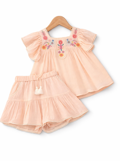 ピーチベージュの女の子用2点セット。花刺繍入りのフリル袖ブラウスと、レーストリム付きのティアードショートパンツ（ウエストゴム・タッセル紐付き）。
