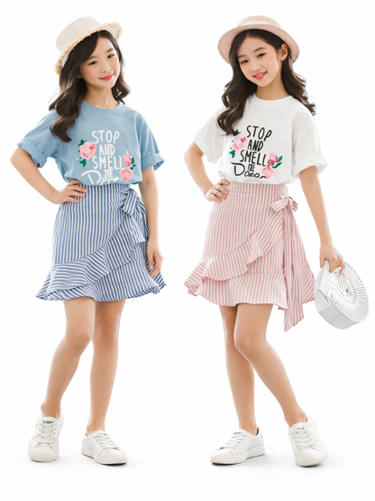 白背景でポーズをとる女の子2人のキッズコーデ画像。ブルーとホワイトの花柄プリント半袖Tシャツに、ブルー／ピンクのストライプフリルスカートを合わせた色違いセット（帽子・スニーカー着用）。