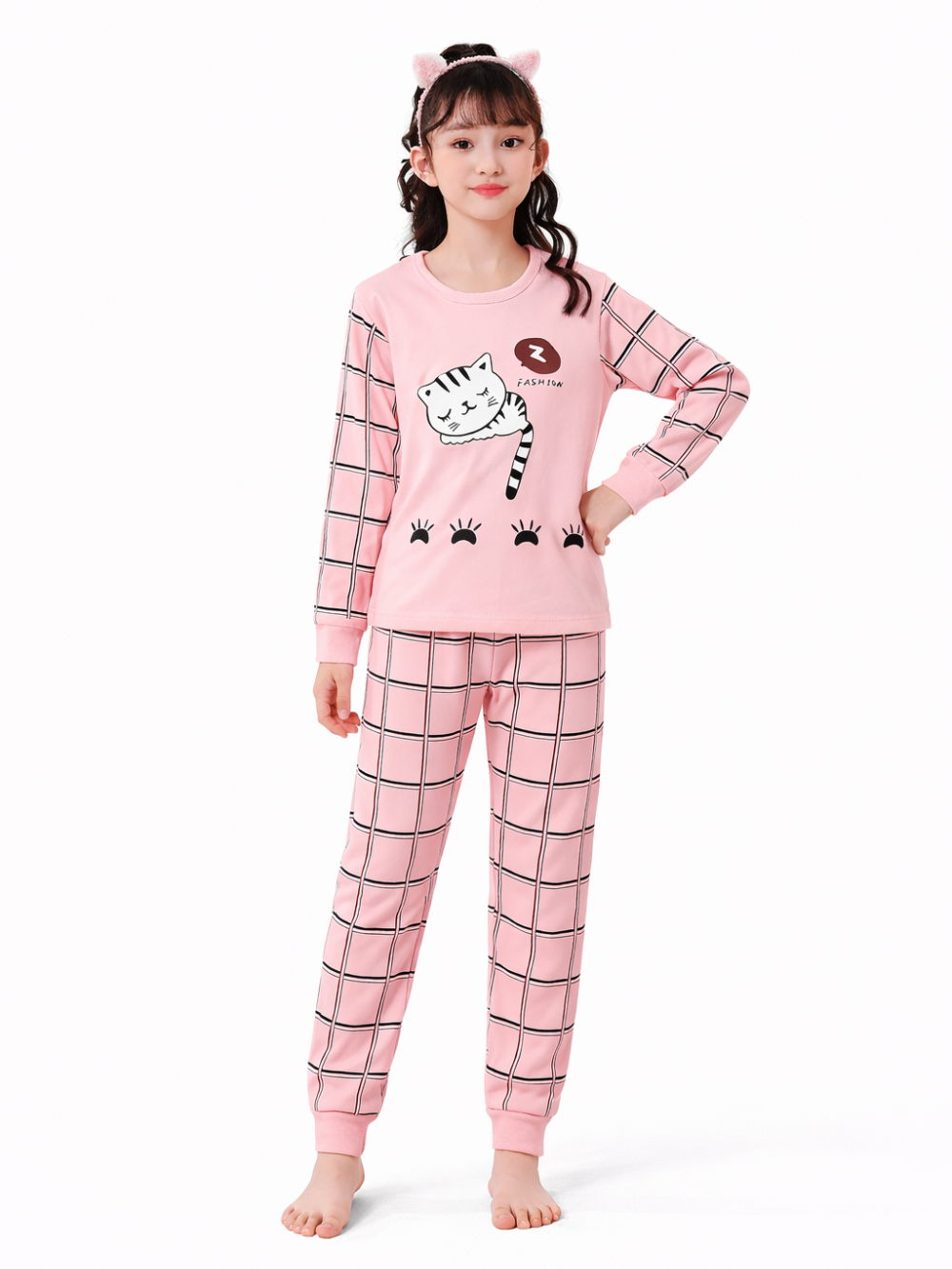 白背景で子ども用ピンクの長袖パジャマ上下セットを着用した女の子モデルの全身写真。猫プリントトップス、チェック柄パンツ、猫耳ヘアバンド付き。