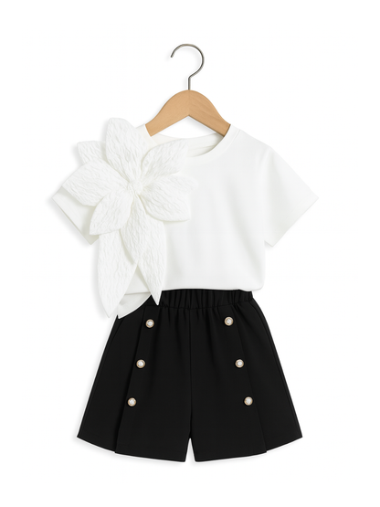 女の子用セットアップ：立体的な白い花の装飾が付いた白の半袖トップスと、パールボタンがアクセントの黒いショートパンツ。スタジオ風の白背景で撮影された高解像度画像。上品で洗練されたデザインが特徴。
