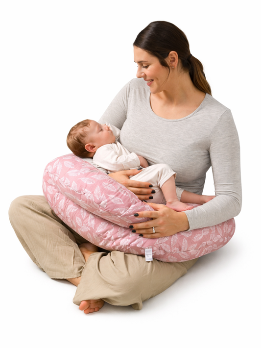 白背景の授乳クッション商品イメージ。ピンクのリーフ柄授乳クッションに赤ちゃんを乗せ、母親が床に座って抱っこしながら授乳している様子を写した写真。
