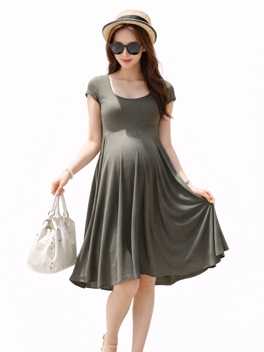 白背景のマタニティワンピース着用イメージ。麦わら帽子と黒サングラスの日本人妊婦モデルが、オリーブグリーンの半袖フレアワンピースを着用し、白いバッグとサンダルを合わせて立ち姿でスカート裾を軽く持ち上げている。
