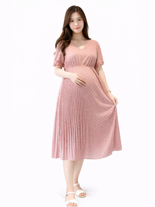 白背景のマタニティワンピース着用イメージ。日本人妊婦モデルが、白ドット柄のピンクミディ丈ワンピース（Vネック・半袖・ウエストギャザー・プリーツスカート）をカタログ風ポーズで着用し、柔らかな表情で立っている。
