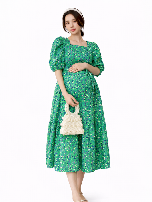 白背景のマタニティワンピース着用イメージ。日本人妊婦モデルが、グリーンの小花柄ミディ丈ワンピース（スクエアネック・パフスリーブ・ウエストリボン・ティアードスカート）を着用し、お腹に手を添えて立ち姿で見せている。
