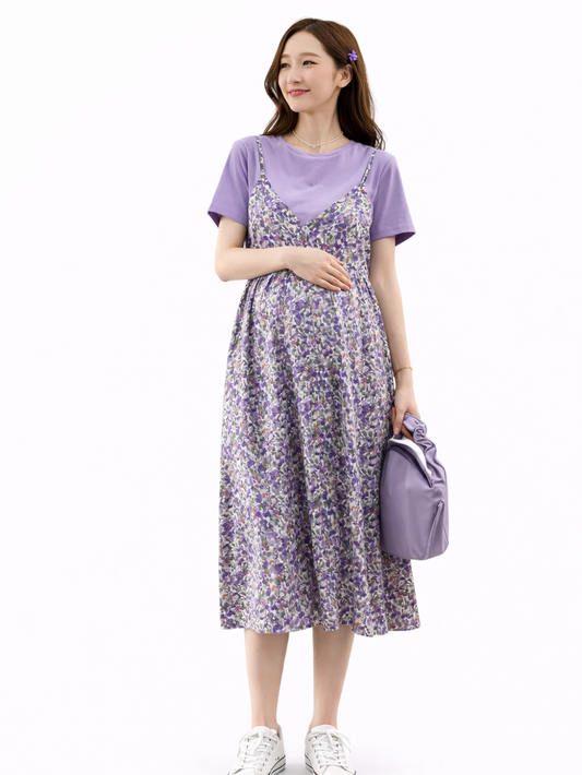 白背景のマタニティワンピース着用イメージ。日本人妊婦モデルが、パープルのTシャツに小花柄のキャミワンピースを重ねたミディ丈ワンピースを着用し、お腹に手を添えて立ち姿でシルエットを見せている。
