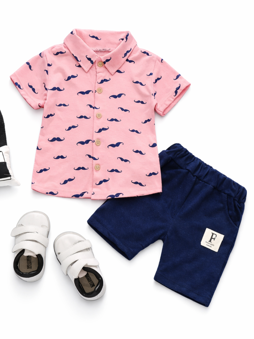 白背景のフラットレイで撮影されたキッズ2点セット。ネイビーのひげ柄プリントが入ったピンクの半袖ボタンシャツと、ウエストゴム仕様のネイビーショートパンツ（小さなタグ付き）。
