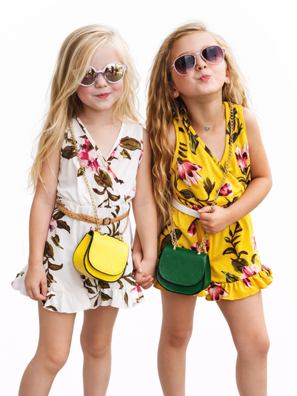 白背景のスタジオ撮影。トロピカルフラワー柄のノースリーブワンピースを着た女の子2人が手をつないで並び、サングラスをかけてポーズ。片方は白地に花柄のワンピース＋黄色のミニショルダーバッグ、もう片方は黄色地に花柄のワンピース＋緑のミニショルダーバッグ。
