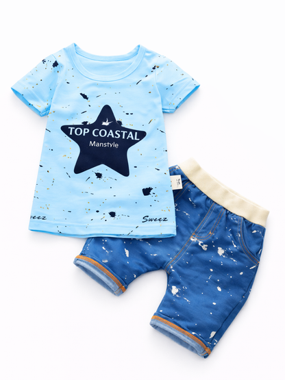 白背景のフラットレイで撮影されたキッズ2点セット。ライトブルーの半袖Tシャツにネイビーの星モチーフとロゴ、スプラッシュ柄プリント入り。クリーム色のリブウエストが付いたブルーのショートパンツは白いスプラッシュ柄とオレンジのステッチが特徴。
