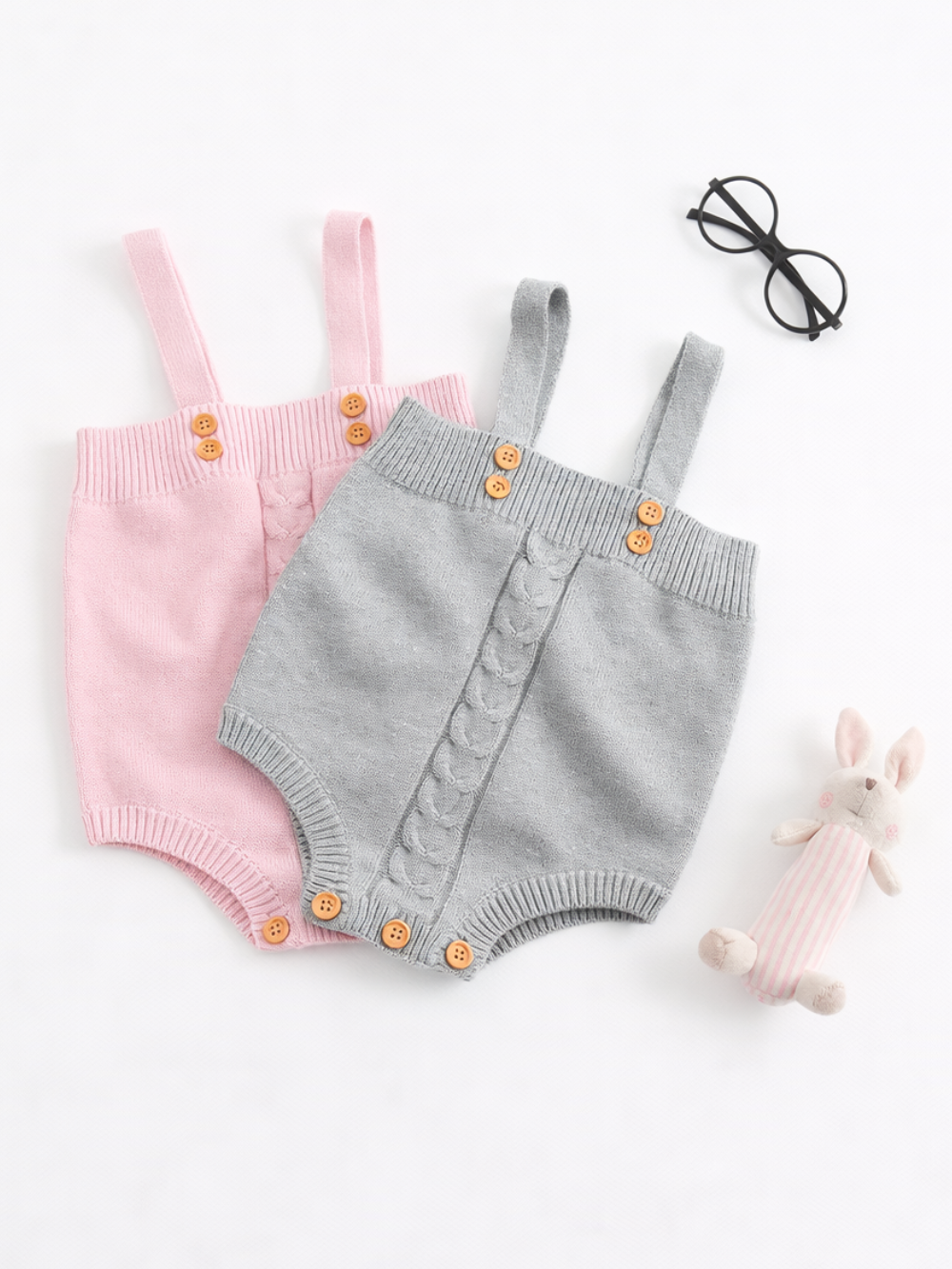 白背景のフラットレイで撮影されたニットベビーロンパース2枚。ピンクとライトグレーのサロペット型で、肩ひもと木製ボタン、リブ編みのウエストと裾、股下スナップボタン付き。近くに黒い丸メガネとピンクストライプのうさぎぬいぐるみが置かれている。
