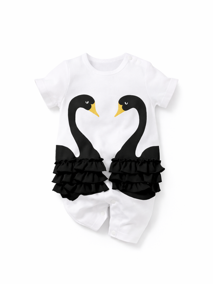 白背景のスタジオ撮影。白い半袖ベビーロンパース単体が中央に配置され、前面に黒い2羽の白鳥プリント（首がハート形）と黄色いくちばしが描かれている。腰まわりには黒いフリルが左右対称に付いており、股下はスナップボタン留め。

